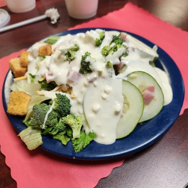 A salad with lettuce, broccoli, cucumbers, ham, croutons, and creamy dressing on a blue plate.