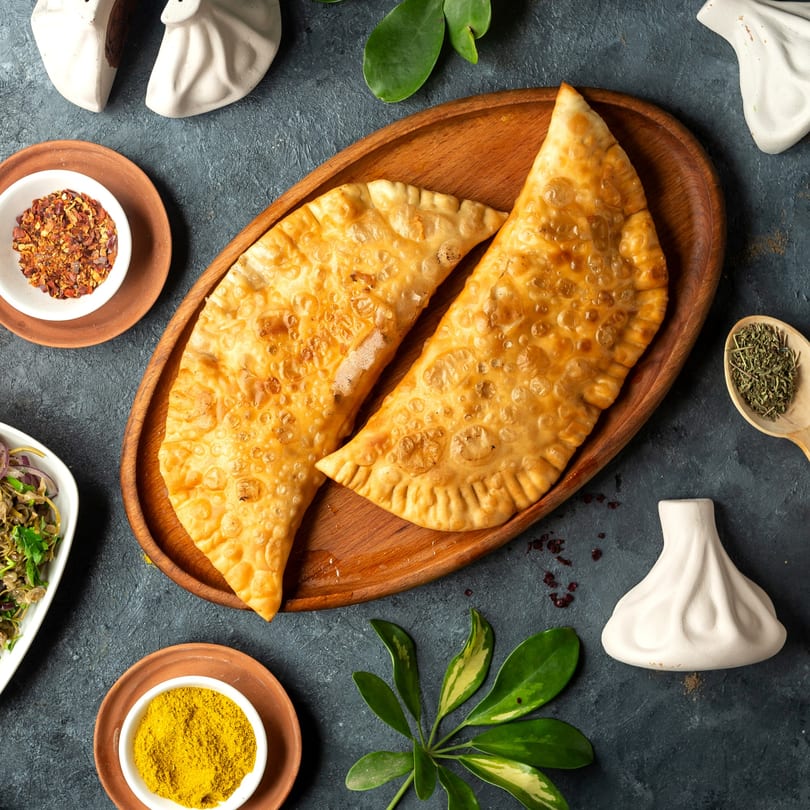 Two golden-brown fried pastries on a wooden tray, surrounded by spices and decorative elements.
