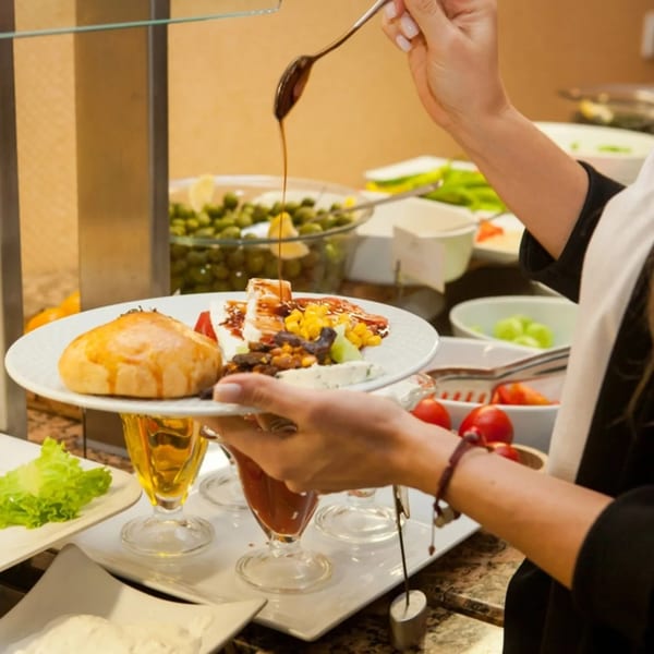 A person's hands holding a plate of food, drizzling sauce over a pastry and salad at a buffet.